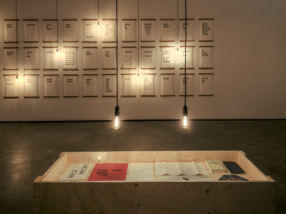 An installation with papers with writing on the wall and two lamps hanging from the ceiling, illuminating a surface with more texts.